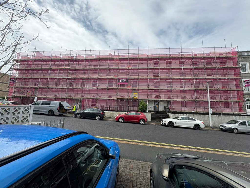 A large building covered in pink scaffolding stands on a sloped street with several parked cars in front. The sky is cloudy, and a "Scaffolding" company sign is visible on the structure.